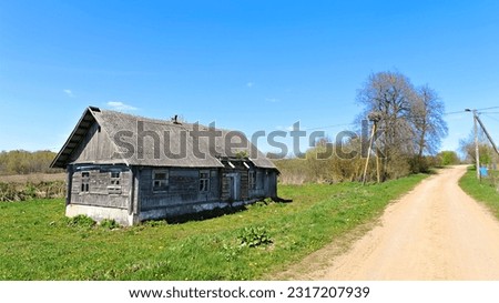 On a grassy lawn stands an old wooden house with a slate roof. Nearby there are trees and bushes. Behind the house is an agricultural field A dirt road and a power line run along the side of the house Royalty-Free Stock Photo #2317207939