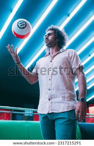 From below shot of focused handsome man in casual outfit throwing up ball with one hand while standing in bowling club.