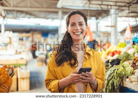 Woman at the farmer's market shopping, sending a text message on her smart phone. Young cheerful woman at the market. Smiling girl decided to cook a delicious and healthy meal Royalty-Free Stock Photo #2317228901