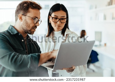 Design professionals standing in an office and using a laptop together. Two young business people discussing a project. Royalty-Free Stock Photo #2317231987