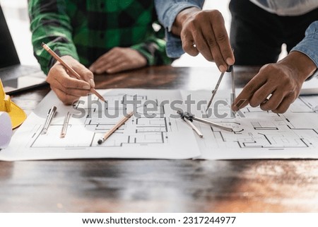 Two engineers wearing yellow clothes working and discussing white grand plan document Two colleagues discussing data and laptop with architectural project at construction site at desk in office Royalty-Free Stock Photo #2317244977
