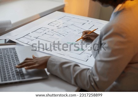 Female architect working with blueprints, cadastral maps and city plans. Young African American lady sitting at her office desk, looking at cadastral maps, studying information and using her laptop Royalty-Free Stock Photo #2317258899