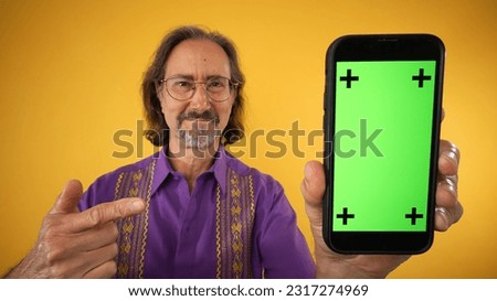 Happy mature elderly hispanic man wear ethnic shirt hold in hand point finger on mobile cell phone with blank green screen workspace area tracking points on solid yellow background studio portrait
