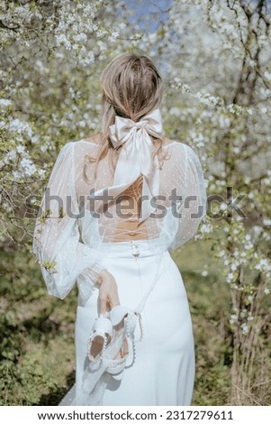 Young woman wearing wedding dress with strings on back standing among cherry trees and holding pair of white high-heeled shoes with hand. Rear view of blonde bride with big bow in hair. Vertical. Royalty-Free Stock Photo #2317279611