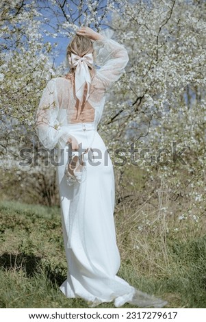 Rear view of young lady with bow in hair wearing long wedding gown with strings on back, standing among cherry blossom and holding pair of white high heels shoes decorated with pearls. Vertical. Royalty-Free Stock Photo #2317279617