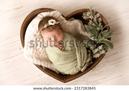A cute newborn baby in a green pistachio winding and a white bandage with a flower on his head sleeps sweetly. Wooden basket in the shape of heart. Professional macro photo against light wooden floor.