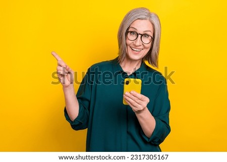 Photo of astonished elegant grandmother hold smart phone indicate finger empty space isolated on yellow color background