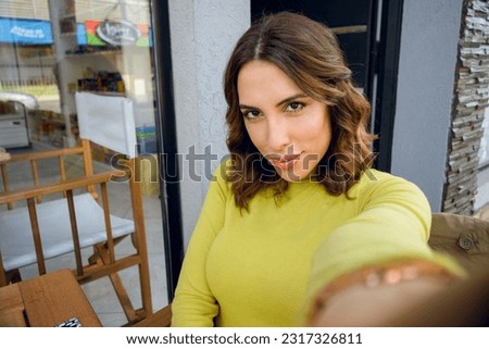 young latina woman of argentinian ethnicity, taking a picture with her phone taking a selfie outdoors looking at the camera, while waiting for breakfast.