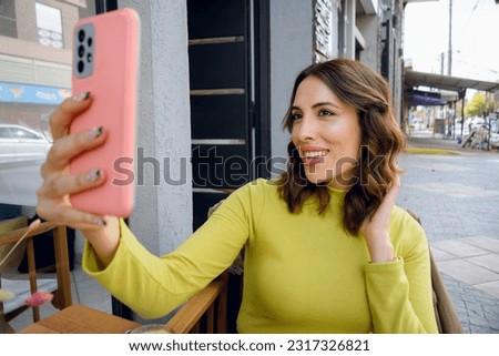 young long-haired latin woman of argentinian ethnicity dressed in a sweater, happy smiling taking a selfie with her phone, sitting outside the coffee shop, technology concept, copy space.