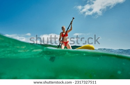 Tanned woman in red bikini sitting on surfboard with paddle relaxing at sea resort enjoying summer