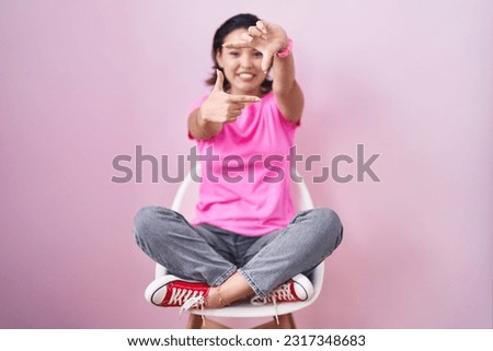 Hispanic young woman sitting on chair over pink background smiling making frame with hands and fingers with happy face. creativity and photography concept. 