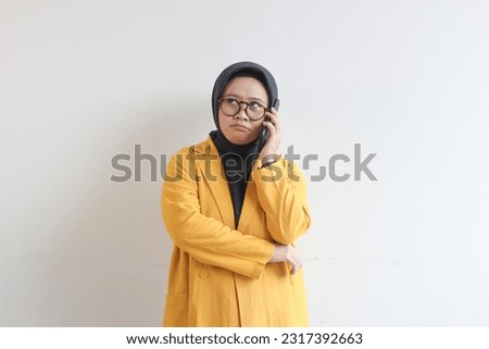 portrait of beautiful asian woman in hijab, glasses and wearing yellow blazer making phone call while looking sideways isolated white background.
