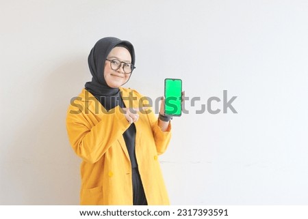 beautiful young asian woman in glasses, hijab and wearing yellow blazer is holding green screen mobile phone with happy face isolated on white background.