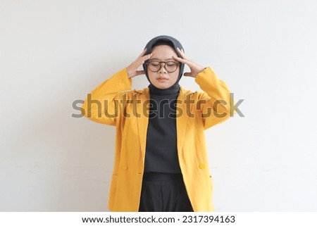 beautiful young Asian Muslim woman, wearing glasses and yellow blazer with dizzy expression holding head isolated on white background.