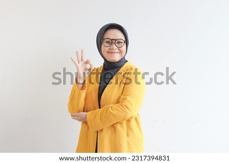 beautiful young Asian Muslim woman, wearing glasses and yellow blazer with hand gesture of approval or OK isolated white background.