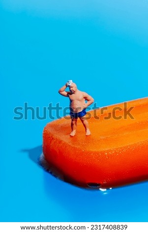 a miniature man, wearing a blue swimsuit, is drying the sweat of his foreground standing on a melting orange popsicle, on a blue background with some blank space on top Royalty-Free Stock Photo #2317408839