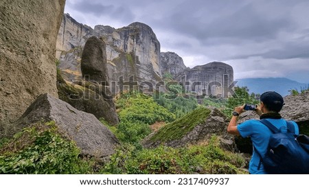 Active man with backpack standing under massive rock formation pinnacles near village Kastraki on mystical hiking trail in Kalambaka, Meteora, Thessaly district, Greece, Europe. Rock climbing activity