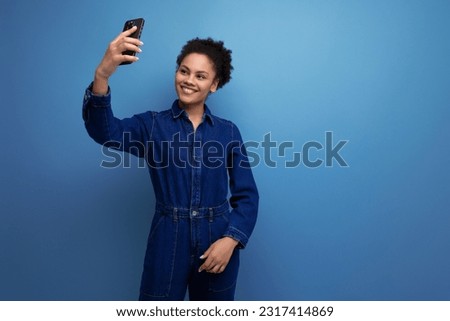 a young woman with dark skin and black curly hair dressed in a blue denim jumpsuit takes a selfie on the phone