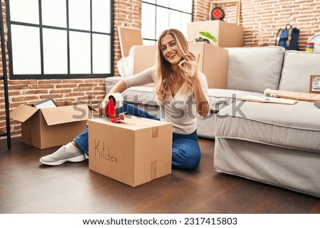 Young blonde woman sitting on the floor closing boxes home doing ok sign with fingers, smiling friendly gesturing excellent symbol 