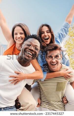 Vertical portrait of two multiracial young friends giving piggyback ride to his girlfriends. A group of happy people having fun and smiling. Men carrying women on his shoulders looking at camera. High Royalty-Free Stock Photo #2317445327