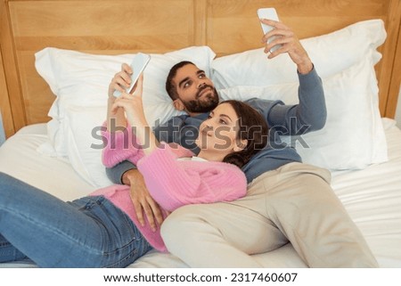 Mobile Technology And People. Young Married Couple Using Smartphones Surfing Web, Texting In Social Media And Having Fun Online Lying In Bed At Home. Modern Gadgets Lifestyle. Selective Focus