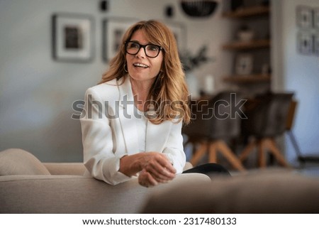 Close-up of a happy attractive woman relaxing in an armchair at home. Blond haired female wearing eyeglasses and white blazer. Royalty-Free Stock Photo #2317480133