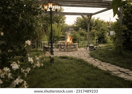 Patio and fire in the fire pit in the evening. Pleasant summer evening. A beautiful fire flame, a glowing lantern and a garland against the backdrop of an evening garden.  Royalty-Free Stock Photo #2317489233
