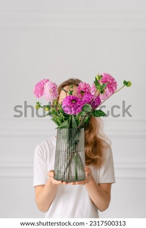 Vertical image of a young woman holding a bouquet of pink dahlias in a vase in front of her on a white background. The concept of flower workshops and articles for the study of floristry Royalty-Free Stock Photo #2317500313
