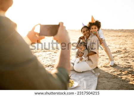 Happy family resting by seaside at sunset, man taking photo of his wife and children on cellphone, male capturing moments of summer day evening