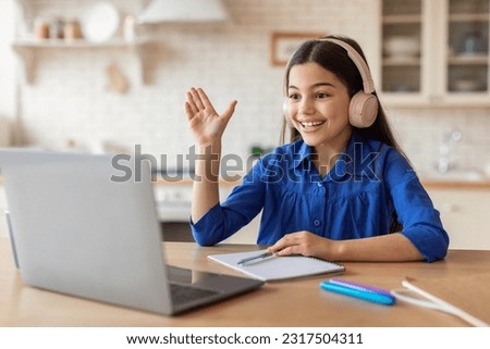 Online Learning Excellence. Schoolgirl With Headphones Raising Hand During Video Call Lesson, Learning Remotely Via Laptop Computer Sitting At Desk At Home. E-Learning Success Concept