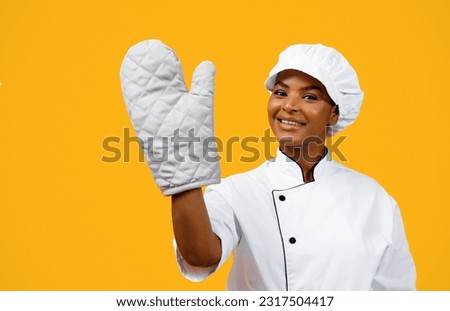 Beautiful Black Female Chef Wearing Oven Mitt Posing Over Yellow Background, Cheerful African American Cooker Lady In Heat Protective Glove And Toque Looking And Smiling At Camera, Copy Space Royalty-Free Stock Photo #2317504417