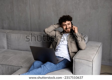 Young handsome millennial man sitting on couch in living room at home using computer chatting and holding smartphone. Laughing positive male talking by cellular with friend feels happy and satisfied