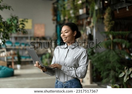 Cheerful young black businesswoman floral store owner standing with laptop in hand with different green plants on background. Female florist working using computer
