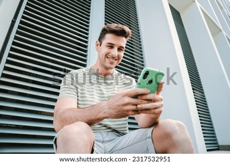 Young guy with friendly expression smiling and having fun playing a video game with his cellphone sitting outdoors. Caucasian teenage stundent enjoying using a mobile phone for watch the social media Royalty-Free Stock Photo #2317532951