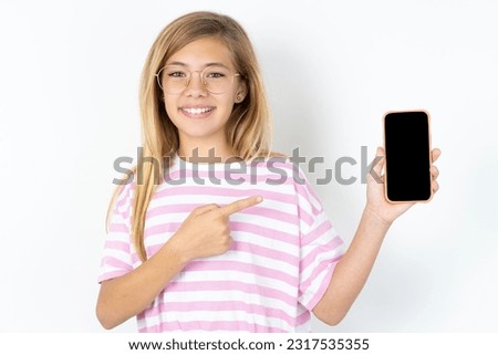 beautiful caucasian teen girl wearing striped T-shirt over white wall looks with bugged eyes, holds modern smart phone, receives unexpected message from friend, reads reminder.