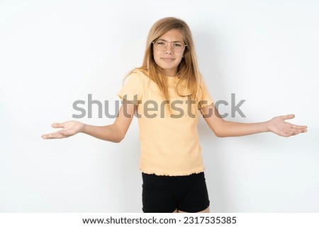 beautiful blonde teen girl wearing orange T-shirt over white studio wall looks uncertain shrugs shoulders.