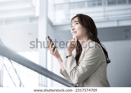 A smiling Japanese woman in a suit and jacket holds a smartphone in a spacious office. Royalty-Free Stock Photo #2317574709