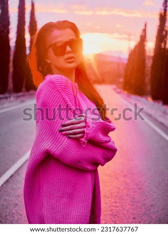 Girl in a pink sweater and sunglasses posing on a mountain road during a warm and cozy sunset Royalty-Free Stock Photo #2317637767