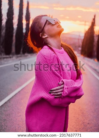 Girl in a pink sweater and sunglasses posing on a mountain road during a warm and cozy sunset Royalty-Free Stock Photo #2317637771