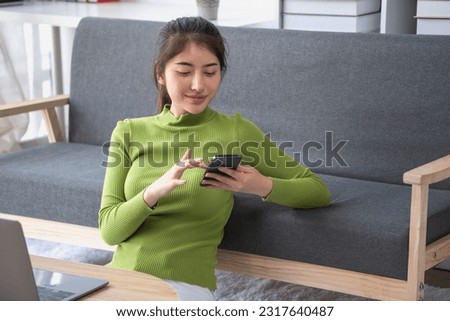 Asian woman with a beautiful face in a green long-sleeve shirt sitting and relaxing watching movies Listening to music on a laptop, mobile phone, and headphones happily inside living room at home.
