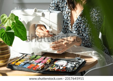 A woman sews tulle on an electric sewing machine in a white modern interior of a house with large windows, house plants. Comfort in the house, a housewife's hobby Royalty-Free Stock Photo #2317645911