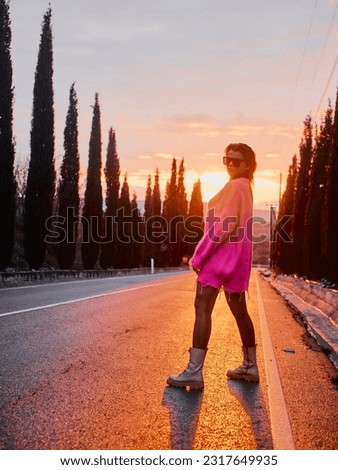 A girl in a pink sweater and sunglasses poses on a car-free road, with a beautiful sunset and a charming alley of Cyprus cypress trees in the background Royalty-Free Stock Photo #2317649935