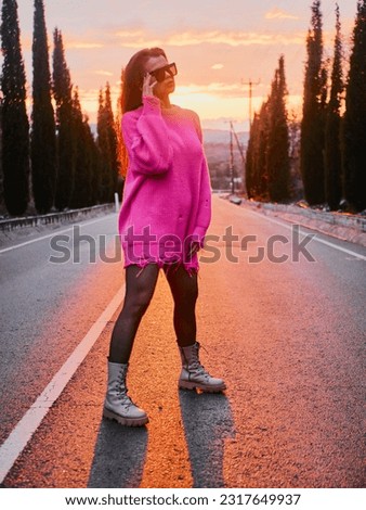 A girl in a pink sweater and sunglasses poses on a car-free road, with a beautiful sunset and a charming alley of Cyprus cypress trees in the background Royalty-Free Stock Photo #2317649937