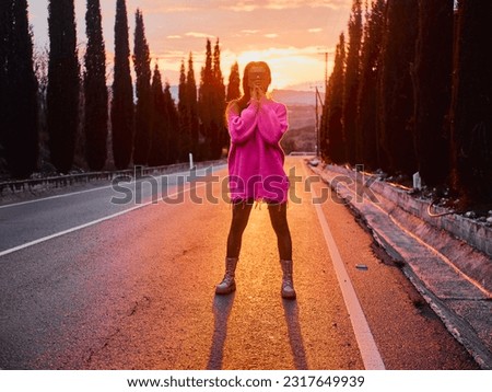 A girl in a pink sweater and sunglasses poses on a car-free road, with a beautiful sunset and a charming alley of Cyprus cypress trees in the background Royalty-Free Stock Photo #2317649939