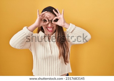 Young hispanic woman standing over yellow background doing ok gesture like binoculars sticking tongue out, eyes looking through fingers. crazy expression. 