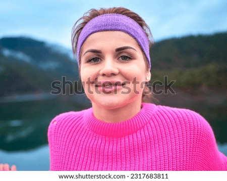 A portrait of a girl in a pink sweater with a sports bandage on her head standing on a dam, set against the backdrop of a majestic mountain Royalty-Free Stock Photo #2317683811