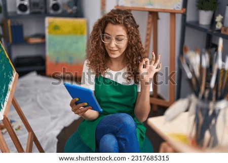 Young caucasian woman painter sitting at art studio doing video call with tablet doing ok sign with fingers, smiling friendly gesturing excellent symbol 