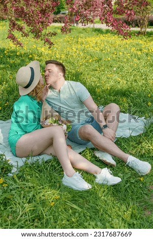 A guy and a girl sit on a picnic blanket in a park, kissing and enjoying their time together. The picture depicts the concepts of weekend, relaxation, dating, and healthy relationship psychology.