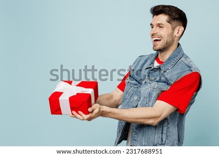 Young fun man he wear denim vest red t-shirt casual clothes hold in hand giving present box with gift ribbon bow isolated on plain pastel light blue cyan background studio portrait. Lifestyle concept
