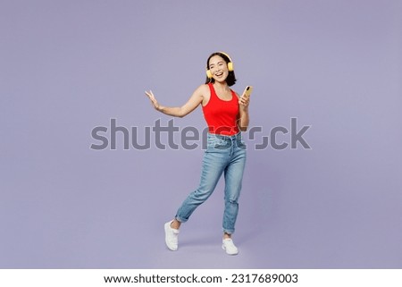 Full body young woman of Asian ethnicity she wears casual clothes red tank shirt headohones listen to music use mobile cell phone isolated on plain pastel light purple background. Lifestyle concept